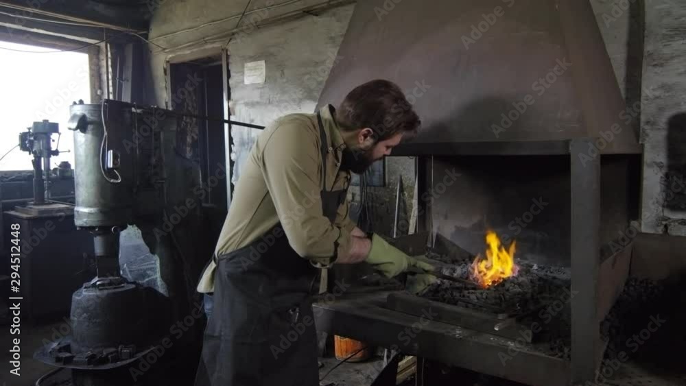 Pan shot of Caucasian bearded blacksmith in apron and gloves approaching to stove with metal billet in his hands, putting it in coals and moving stove damper to blow fire up Pan shot of Caucasian bearded blacksmith in apron and gloves approaching to stove with metal billet in his hands, putting it in coals and moving stove damper to blow fire up