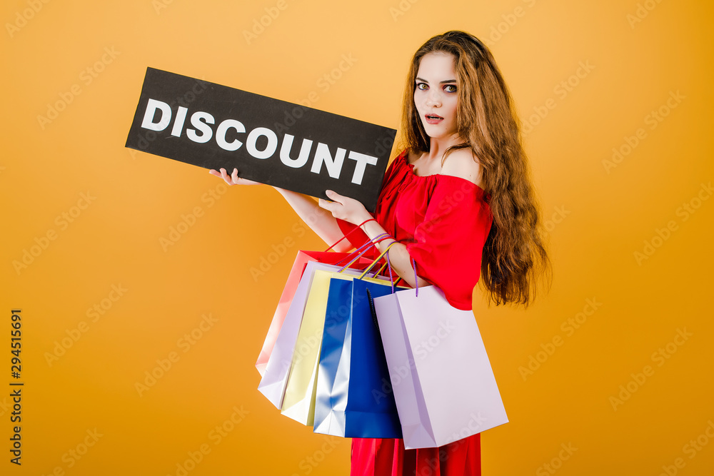 young woman in red dress with discount sign and colorful shopping bags isolated over yellow young woman in red dress with discount sign and colorful shopping bags isolated over yellow