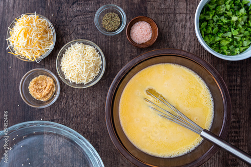 Ingredients for quinoa egg bake, raw egg mix in glass bowl with metal whisk, quinoa, cheese, herbs, broccoli, glass baking dish, on a wood table photo