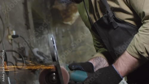 Tilt down of bearded Caucasian craftsman in safety apron, gloves and goggles standing at workbench in smithy with electrical grinding machine in his hands and polishing iron detail photo