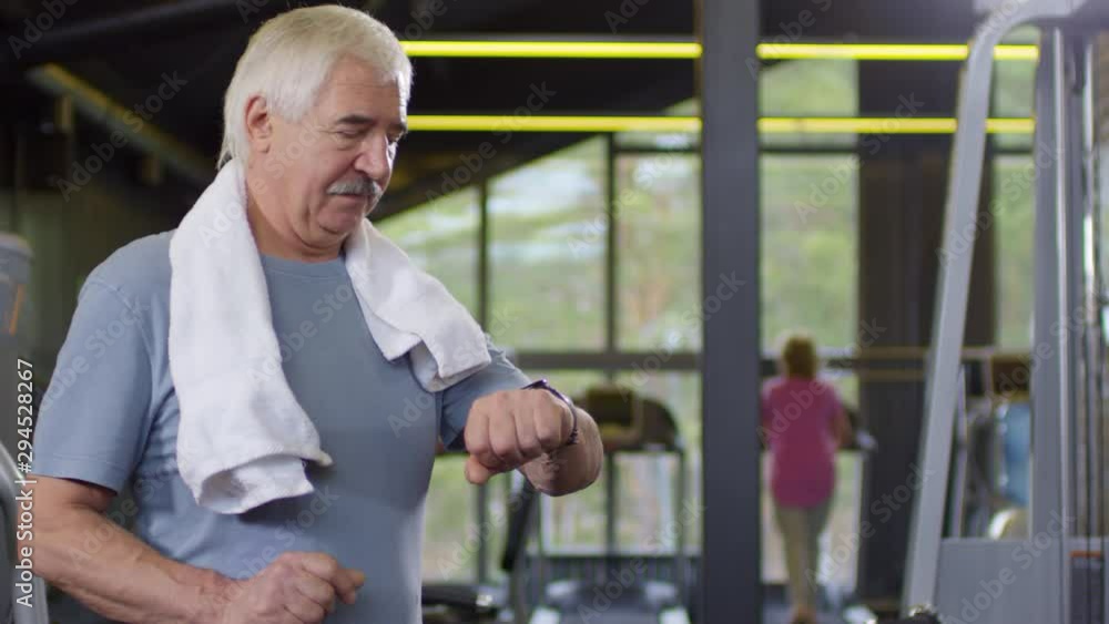 Tracking left of aged Caucasian man in sport clothes standing with towel on his shoulders and checking his smart watch Tracking left of aged Caucasian man in sport clothes standing with towel on his shoulders and checking his smart watch