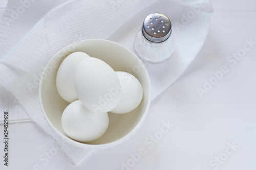 four white eggs lie in a plate on a white wooden table next to a towel and salt shaker.