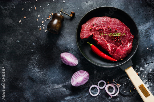 Raw beef steak with spices, onions and chili in cast-iron frying pan for frying on dark slate or concrete background. Top view.