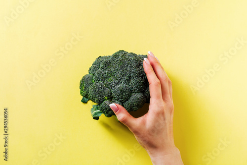 Female hand holds one fresh green broccoli lying on yellow light background. Top view.