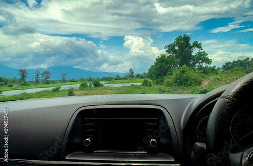 Beautiful view from inside car. Steering wheel and dashboard of car interior. Road trip travel with scenic view of mountain, lake, and forest. Blue sky and white fluffy clouds. Vacation time.