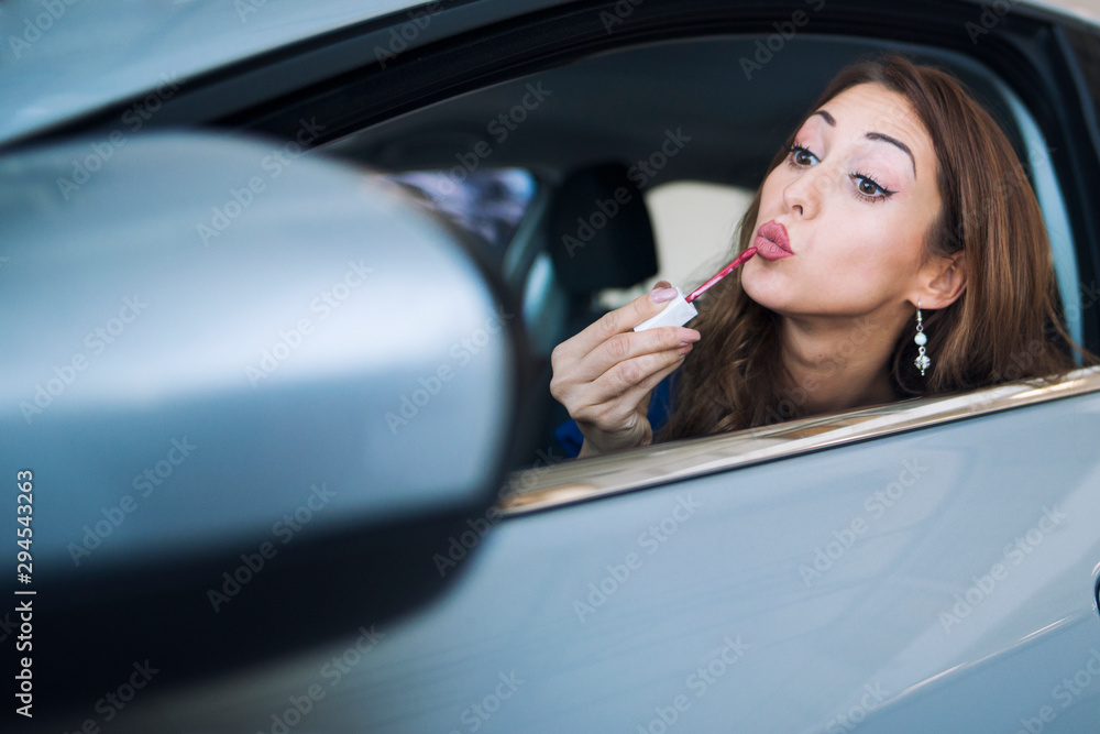 Shot of pretty woman driver siting in her car looking at rear view mirror and putting lipstick on and applying make up.