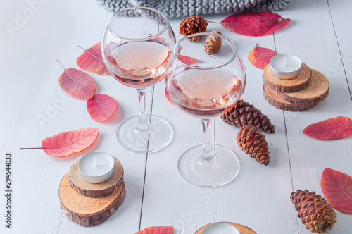 Small candles, two glasses with rose wine, cones, dry red leaves, gray scarf knitted on a white wooden table. Hello, Autumn. Cozy autumn background.