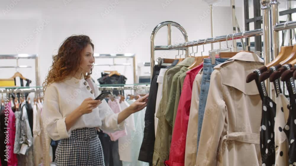 Medium shot of Caucasian woman with long curly hair with smartphone in her hand choosing clothes in shop Medium shot of Caucasian woman with long curly hair with smartphone in her hand choosing clothes in shop
