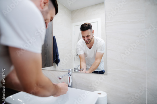 Handsome caucasian man in pajamas washing his face in washbasin in morning. Bathroom interior.