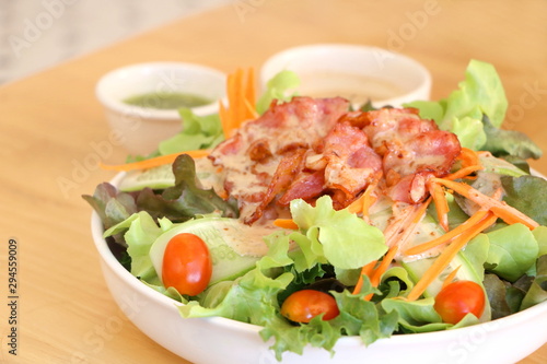 Close up fresh vegetable salad (lettuce, carrot, cucumber, tomato) with fried bacon and roasted sesame Japanese dressing in white bowl on wooden table background