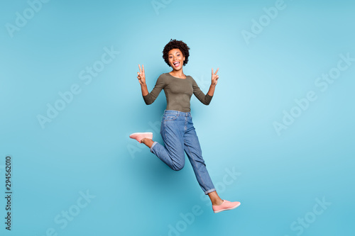 Full length body size photo of cheerful funny screaming friendly nice dark skinned woman jumping jeans denim green sweater showing you v-sign isolated over vivid color blue background running