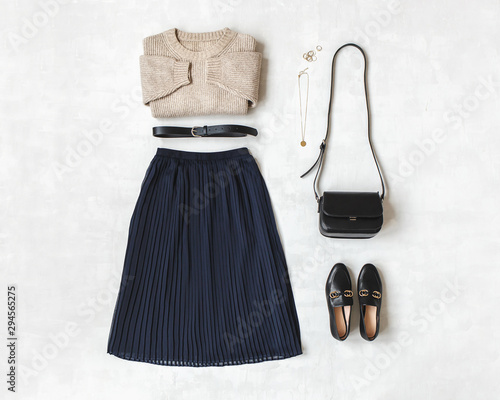 Blue midi pleated skirt, beige knitted sweater, small black cross body bag, belt, loafers (flat shoes) on grey background. Overhead view of women's casual day outfit. Flat lay, top view. Women clothes photo