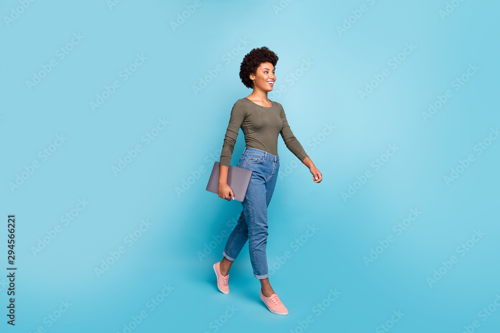 Full length photo of positive cheerful afro american girl hold her netbook go university for education learning wear casual style outfit denim jeans pink sneakers isolated blue color background Full length photo of positive cheerful afro american girl hold her netbook go university for education learning wear casual style outfit denim jeans pink sneakers isolated blue color background