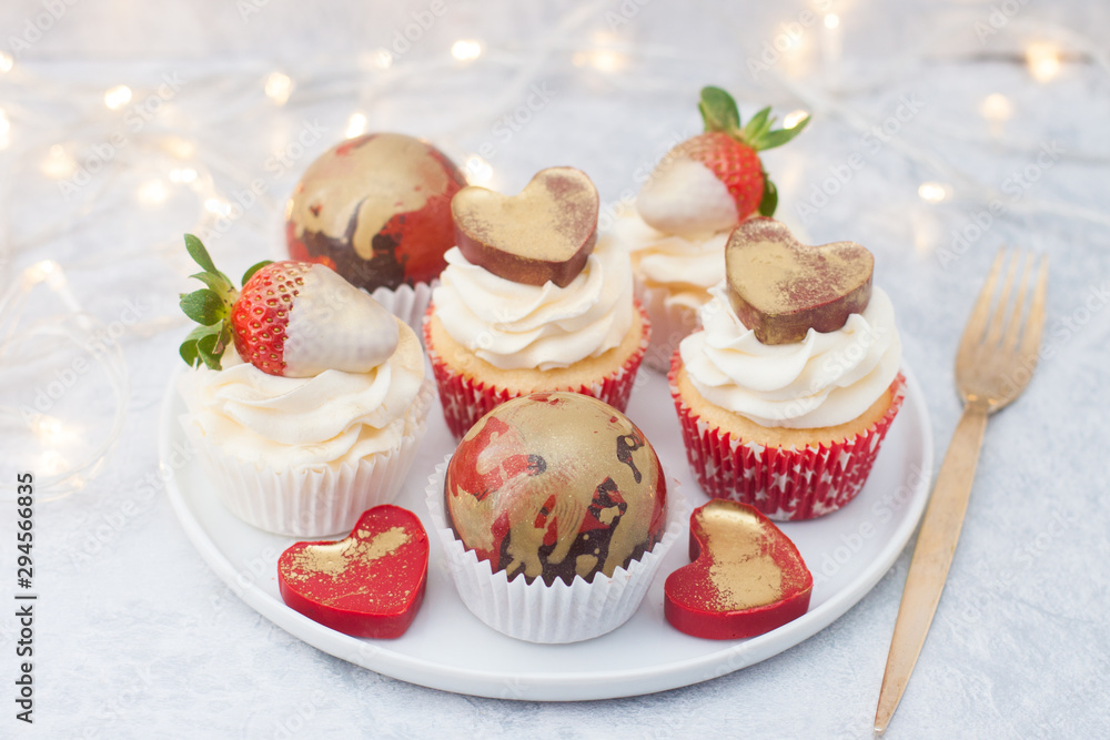 Cupcakes with cream cheese frosting decorated with fresh strawberries in white chocolate, chocolate red and golden balls and red chocolate hearts on the white plate. String lights on background.