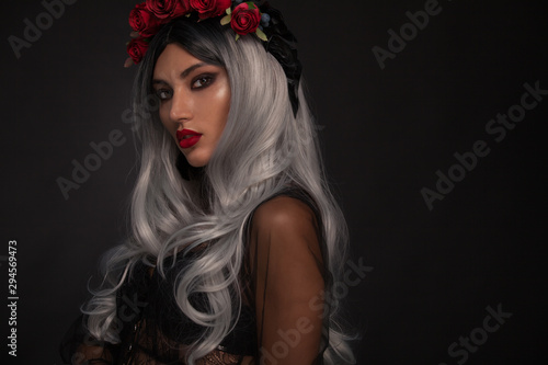 Creative image of Halloween makeup look or Dia De Los Muertos holiday on dark background with copyspace. Blonde model wearing a hat with red rose
