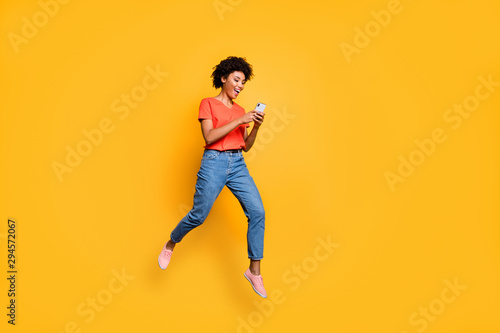 Full length photo of positive cheerful funny curly brown hair girl use smartphone read post blogs in social network jump wear red t-shirt denim jeans isolated over yellow color background © deagreez