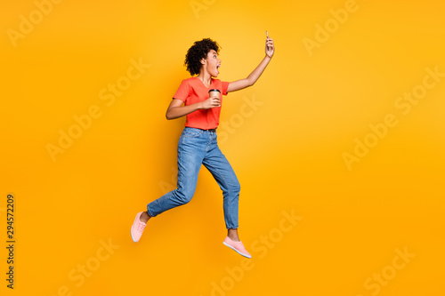 Full body profile side photo of crazy funny wavy hair beautiful girl use cellphone take selfie trip journey hold mug jump wear t-shirt jeans sneakers isolated over yellow color background