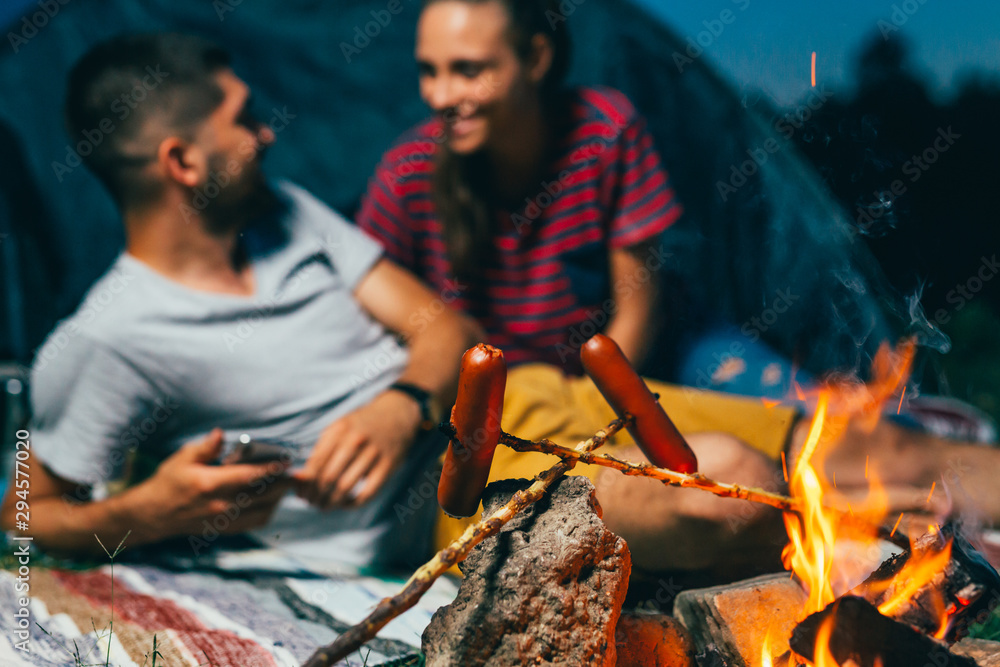 Fototapeta premium close up of campers barbecue sausages. happy young couple on night camping by the camping fire