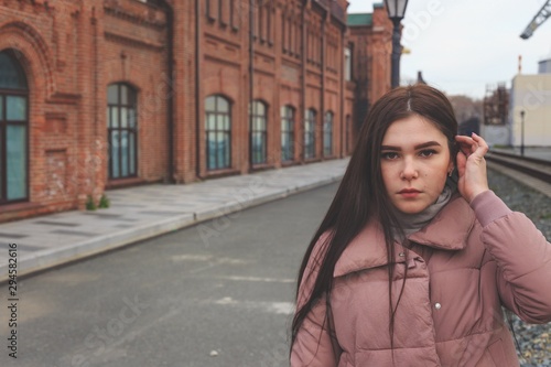 A young pretty pregnant girl in a pink warm down jacket walks near the old railway station. Late autumn, the cold is coming. © Николай Крапивин