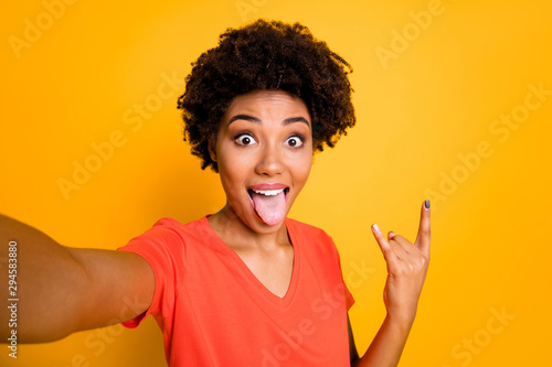 Photo of attractive cheerful sweet pretty curly trendy wavy foolish girlfriend sticking her tongue out taking selfie with fingers horned while isolated over yellow vivid color background