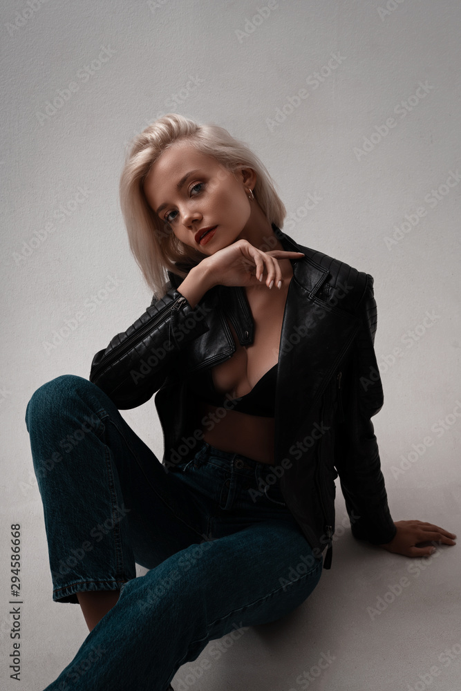 Hot trendy fashion woman model in leather jacket, jeans and underwear posing on white background in room. Portrait of sexy stylish hot blonde girl in lingerie with beauty face and body. Hot trendy fashion woman model in leather jacket, jeans and underwear posing on white background in room. Portrait of sexy stylish hot blonde girl in lingerie with beauty face and body.