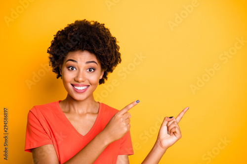 Phoo of curly wavy cheerful cute charming fascinating girlfriend pointing at emptiness with her forefingers smiling toothily wearing orange t-shirt isolated over vivid color yellow background photo