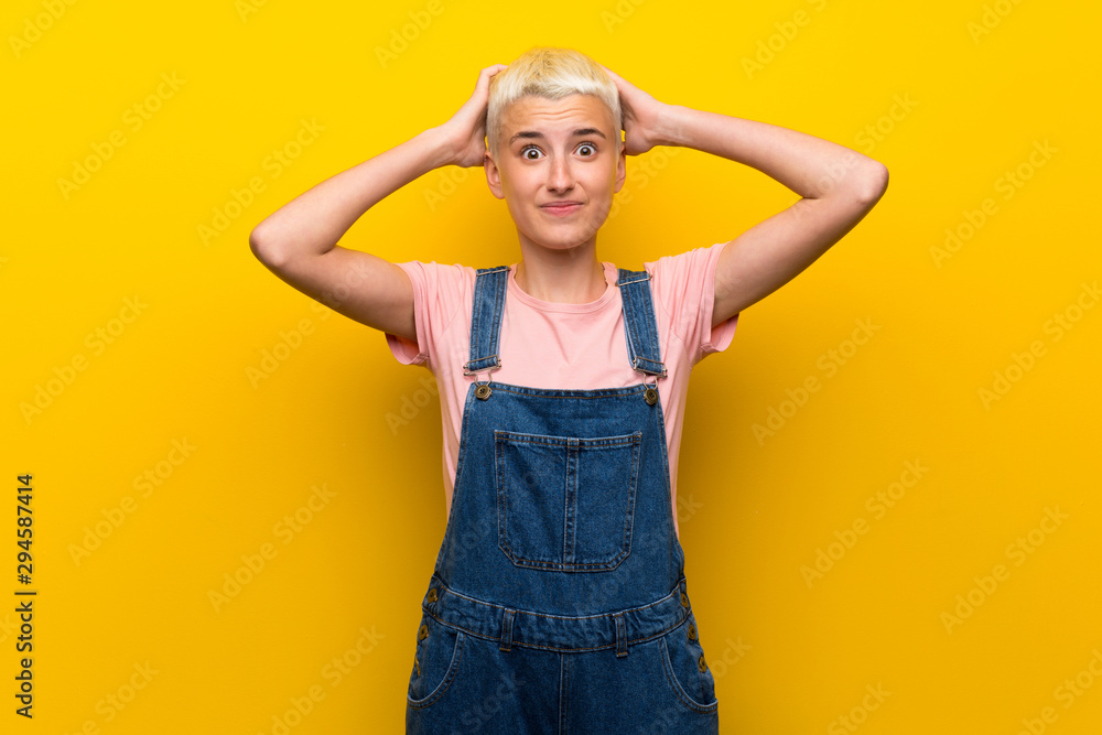 Teenager girl with overalls on yellow background frustrated and takes hands on head Teenager girl with overalls on yellow background frustrated and takes hands on head