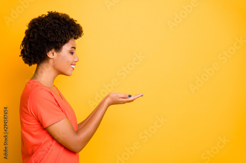 Copyspace side profile photo of cheerful cute charming nice girlfriend holding something with her hands looking at it wearing orange t-shirt isolated over yellow vibrant color background
