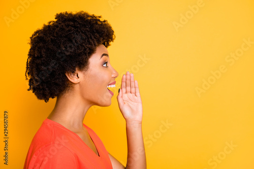 Photo of pretty wavy dark skin lady sharing new information with people holding arm near mouth wear orange t-shirt isolated yellow color background