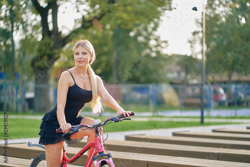 Fototapeta Naklejka Na Ścianę i Meble -  young woman in a short black summer dress rides a bicycle in a city park