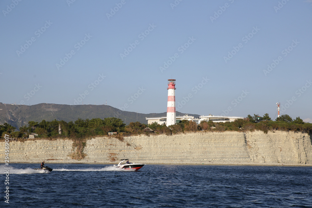 lighthouse on coast of sea. White and red color lighthouse. Against the blue sky. A yacht and a jet ski pass by. View from the sea.