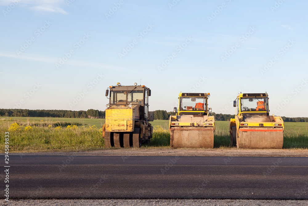 Three large yellow asphalt paving rollers standing on the side of the track during road repair and laying black new asphalt against a landscape with a blue sky and clouds in the summer.