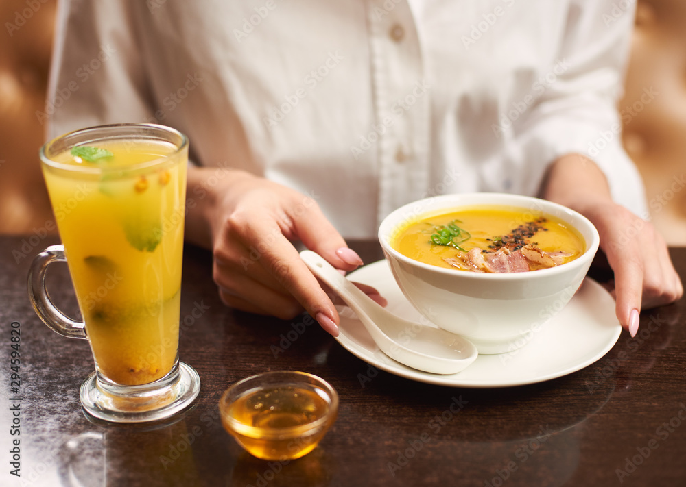 Woman in white blouse ordering tasty and delicious meal at restaurant. Female hands touching white bowl with orange cream soup. Near first dish on brown table standing tea with mint and honey. Woman in white blouse ordering tasty and delicious meal at restaurant. Female hands touching white bowl with orange cream soup. Near first dish on brown table standing tea with mint and honey.