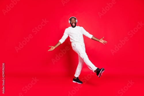 Full body photo of amazing dark skin man listening favorite playlist in earflaps moving with rhythm down street wear knitted sweater trousers isolated red background