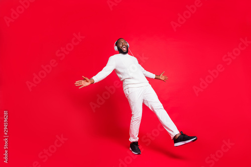 Full length photo of amazing dark skin man listening favorite playlist in earflaps enjoy best moment moving with rhythm wear knitted sweater and trousers isolated red background