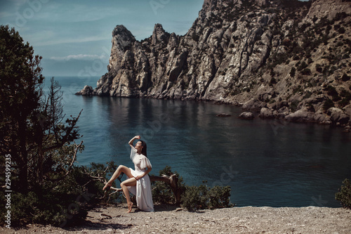 A beautiful girl, a bride, in a white dress sits a tree, on a cliff against the background of the ocean and mountains. With makeup and hairstyle. Fashion, style, wedding. Space.
