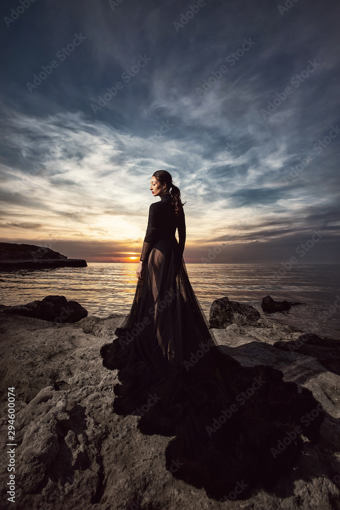 A beautiful girl in a long black dress poses at sunset amid the sea. Beautiful sky. A beautiful girl in a long black dress poses at sunset amid the sea. Beautiful sky.