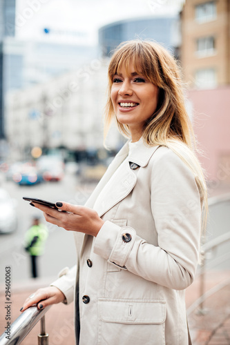 Positive woman with charming smile.beautiful blonde in beige trench coat holding smartphone. against the backdrop of a big city