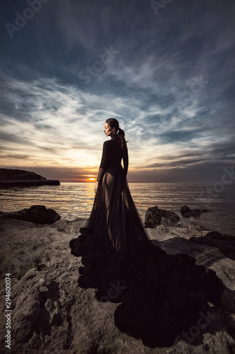 A beautiful girl in a long black dress poses at sunset amid the sea. Beautiful sky.