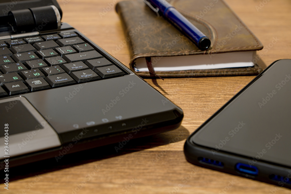 Remote workplace. Remote work environment. View of a natural wood table with several items, a black laptop, a diary, a pen and a mobile phone. Remote workplace. Remote work environment. View of a natural wood table with several items, a black laptop, a diary, a pen and a mobile phone.