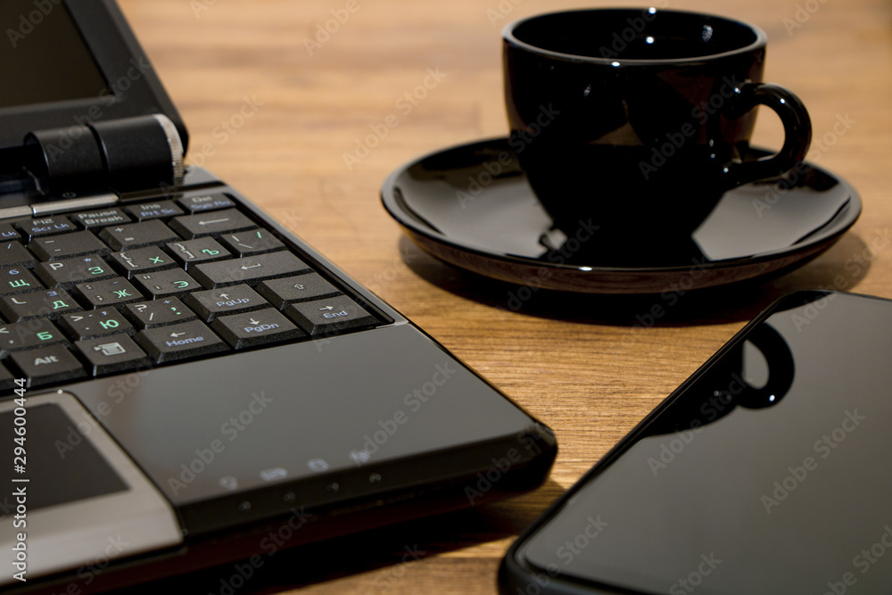 Remote workplace. Remote work environment. View of a natural wood table with several items, a black laptop, a cup of coffee and a mobile phone. Remote workplace. Remote work environment. View of a natural wood table with several items, a black laptop, a cup of coffee and a mobile phone.
