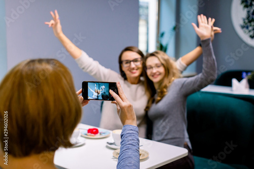 Fototapeta Naklejka Na Ścianę i Meble -  Women friends in cafe indoors. Two pretty women friends hugging, with hands up and posing for the photo together, while her third friend woman is taking a picture on the smartphone