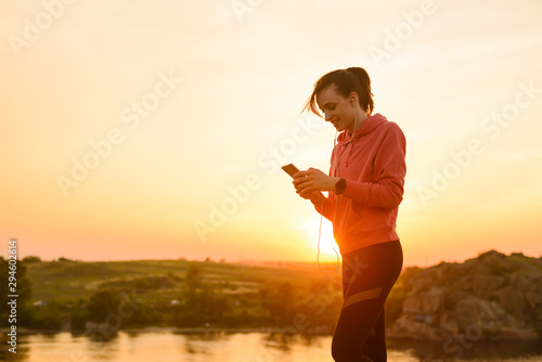 Woman Runner Resting after Workout, Using Smartphone and Listening to Music at Sunset on the Rock. Sports Concept.