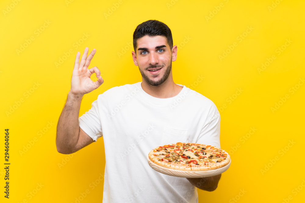 Young handsome man holding a pizza over isolated yellow wall showing ok sign with fingers