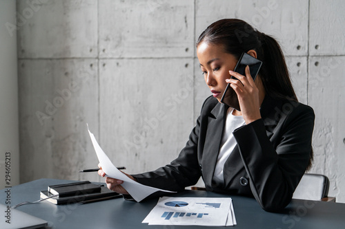 Beautiful young Asian businesswoman talking on smartphone and reading report in blurred loft office. Concept of paperwork and business lifestyle. photo