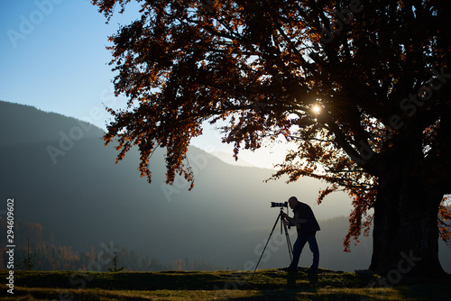Hiker photographer taking picture of misty mountain landscape using professional camera on tripod, on quiet autumn evening standing on grassy valley under big tree under blue sky at sunset.