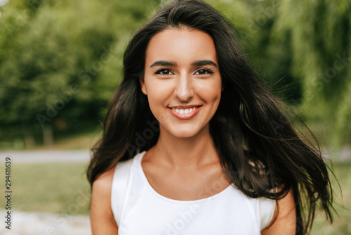 Outdoor close-up portrait of a beautiful young brunette woman smiling broadly and looking to the camera, posing against nature background in the park Fototapete