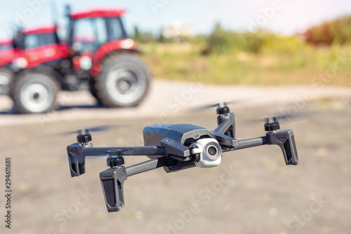 Compact black drone quadcopter with surveillance camera flying low against red agricultural tractor machines. Usage modern technologies and agriculture equipment machinery for farming and agronomy
