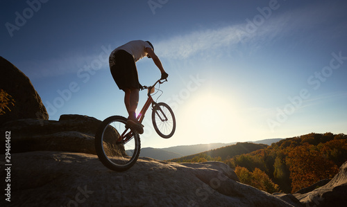 Silhouette of professional sportsman cyclist riding on back wheel on trial bicycle, man rider making acrobatic trick on the big boulder in the summer sunny evening. Blue sky and sunset on background.