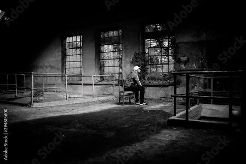 Black and White - Man with white cap and red jacket sitting in front of a broken window in an old and abandoned building